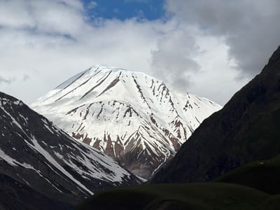 The image captures a dramatic mountain landscape, likely in Gudauri, Georgia, characterized by a towering, snow-capped peak dominating the center. The mountain's slopes are a stark contrast of white snow and dark, exposed rock and earth, with visible streaks of snowmelt creating jagged patterns. The scene is framed by two dark, steep mountainsides, one on the left with patches of snow, and another on the right that is almost entirely in shadow. The sky above is a mix of bright blue and overcast grey clouds, with sunlight intermittently breaking through, illuminating the main peak. In the lower right portion of the image, on a dimly lit grassy slope, a small group of people are gathered around what appears to be a yellow structure, suggesting a point of interest or activity.
