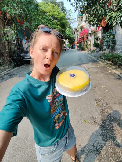 A woman stands in a street in Thành phố Hà Giang, Vietnam, holding a round, yellow cake in a clear plastic container. She wears a green t-shirt with tourist destinations printed on it, denim shorts, and sunglasses. Her mouth is open in surprise or excitement. Behind her, a narrow street extends with buildings, trees, and foliage on either side. A black car is parked near a building on the left, with license plate "23A-109.80." A mixture of sunlight and shade patterns the street surface.