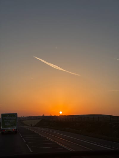 A vertical, low-angle shot shows a road stretching into the distance with a van on the left and a sunset on the right. The van, a white ASDA delivery truck, is positioned in the foreground on the left side of the frame. Its rear is facing the viewer, and text on the back reads "What do you fancy for dinner tonight? Same day delivery now available ASDA.com". The road has multiple lanes marked with white lines.
The right side of the image features a sunset with the sun as a bright orange orb low on the horizon. The sky transitions from a deep blue at the top to a warm orange and yellow near the horizon. A faint contrail from an aircraft is visible in the upper part of the sky. The horizon is lined with a dark silhouette of hills and a wooden fence. The environment appears to be rural, with fields visible behind the fence. The overall lighting suggests either dawn or dusk.
The location is identified as Rottingdean, United Kingdom, likely along a road with views of the South Downs.
