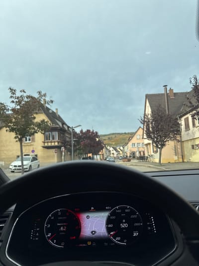 The image depicts a static street scene captured from the driver's perspective inside a stationary vehicle on Rue du Général de Gaulle, Kaysersberg-Vignoble, France, looking generally east-southeast under an overcast sky.
In the immediate foreground, a dark, multi-function steering wheel and a digital instrument cluster are visible. The cluster displays vehicle operational data: current time 16:03, ambient temperature 16.5°C, 0 km/h speed, 0 RPM, D4 gear selection, 6.2 l/100km fuel consumption, 4983.8 km total distance, an active navigation map, and illuminated tire pressure and general warning indicators.
The midground reveals a paved street bordered by multi-story, traditional Alsatian-style buildings. On the left, a distinctive light yellow/ochre building with a brown roof and a prominent wooden-framed bay window/balcony is visible. A small tree with autumn foliage stands in front of this building, with a white BMW 5-series sedan parked roadside. A red circular "No Entry" sign is present further down on the left. On the right, multiple light orange/peach and beige colored buildings with brown roofs line the street, partially obscured by trees with reddish-brown leaves. Outdoor seating areas with tables and chairs are discernible further along the street on the right. Several other vehicles are visible in the distance.
The background features rolling hills covered in vineyards, exhibiting green, yellow, and light brown autumnal colors, rising above the town's architecture. No individuals are distinctly identifiable.