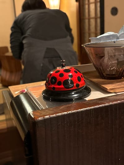 The image shows a red and black ladybug-shaped bell on a dark wooden table. The bell is sitting on a black rectangular base. The table is partially obscured by a dark gray jacket worn by an individual out of focus in the background, standing to the left of the image. There is a glass bowl to the left of the bell, filled with a white substance, partially obscured by the jacket. The table appears to be in a restaurant or cafe setting, based on the presence of the bell. The location, based on the coordinates provided, is within the city of Naoshima, Japan.