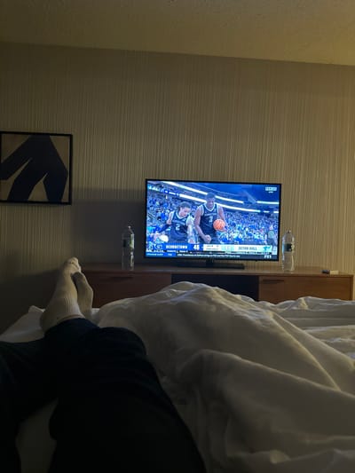 The image captures an intimate, relaxed scene, likely from a bedroom or hotel room, taken from the perspective of someone lounging in bed. The photo is rotated 90 degrees counter-clockwise from its natural orientation, showing a television screen as if it were vertical.

In the center of the frame, a flat-screen television is mounted on a wall featuring horizontal striped wallpaper. The TV is displaying a live basketball game. On the screen, two players in blue jerseys are visible on the court; one player (number 1) holds the basketball, while another player (number 5) is nearby. The scoreboard overlay indicates "SETON HALL 48" versus "GEORGETOWN 46" with "2:24" remaining in the "2ND HALF". A "BIG EAST" logo is prominently displayed, along with smaller text indicating "FOX" and "FS2" as the broadcast channel, and a "Program Alert".

Below the television, on what appears to be a narrow wooden shelf or console, two clear plastic water bottles are placed. Further down the wall, below the TV and the console, a dark-framed piece of art hangs, featuring an abstract blue shape against a lighter background. The wall to the far left of the TV has a different, more granular texture compared to the horizontally striped wallpaper.

To the right side of the image, a large, rumpled white duvet or blanket covers the majority of a bed. From beneath the duvet, the lower legs and feet of a person are visible, clad in white socks. Dark pants or clothing are also partially seen around the ankles. Part of a dark wooden furniture piece, possibly a headboard or nightstand, is discernible behind the white bedding. The overall lighting is subdued, suggesting a comfortable, indoor evening or night setting.