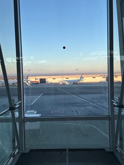 The image displays an airport tarmac scene, viewed through the large windows of a terminal building or jet bridge. The foreground is dominated by dark window frames, metal support structures, and reflective glass, featuring a noticeable red circular reflection. To the right, an interior section shows a metal handrail and a patterned wall or partition with a geometric, zigzag design.

The sky in the background exhibits a soft gradient, transitioning from a pale yellow-orange near the horizon to a clear light blue overhead, indicating either early morning or late afternoon light. The weather appears clear and cloudless.

On the tarmac, multiple aircraft are parked. A prominent white airplane with blue accents is positioned directly beneath the window, seemingly docked with a passenger stair or jet bridge connected. Further in the distance, another aircraft can be seen, partially displaying what appears to be an Air France livery on its tail, characterized by red, white, and blue stripes. The tarmac itself features various white ground markings.

A long, light-colored building runs parallel to the tarmac in the background. On this building, the text "FAST BAGGAGE ITER by ONDA" is clearly visible. Additional airport ground equipment, such as light poles, are also sparsely distributed across the scene.

The image captures a quiet moment of typical airport operations, with aircraft stationed on the apron, likely undergoing pre-flight preparations, servicing, or awaiting passengers. Given the context of Nouaceur, Morocco, the location is likely Mohammed V International Airport (CMN) in Casablanca.