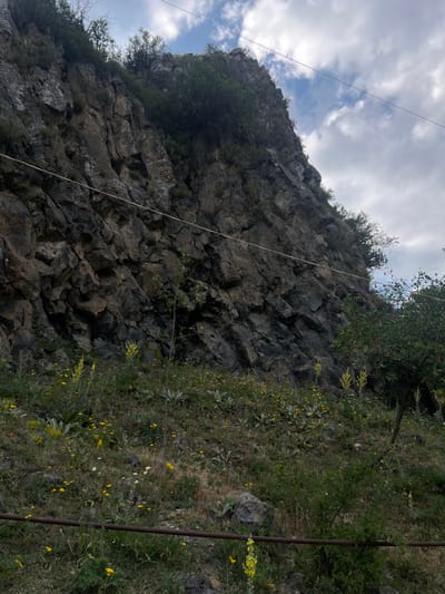 A steep rocky hillside dominates the image, with scattered green vegetation clinging to its surface. The rocks are dark and angular, forming a jagged texture. A thin white cable stretches across the scene, seemingly connecting the hillside to an unseen point. The sky is visible at the top of the frame, filled with wispy clouds. The vegetation appears more abundant on the right side of the image.