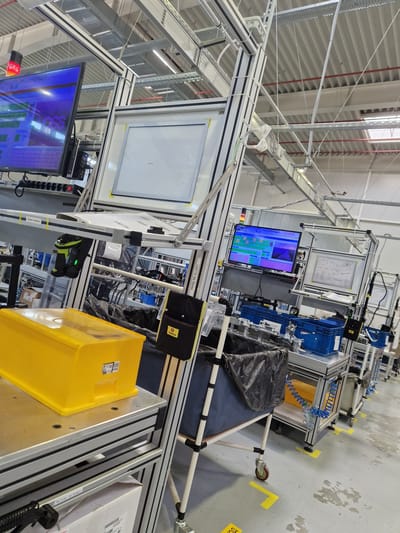 The image depicts an indoor industrial setting, likely a factory or workshop, with a focus on an assembly line. The scene is well-lit with overhead fluorescent lighting and appears to be in a clean, organized environment.
In the foreground, a bright yellow plastic container rests on a metal workbench. To its left, a black and silver barcode scanner with a green light is mounted on a movable arm. Behind this, a large monitor displays a blue interface with text and what appear to be grid-like structures. Further back, a white board with a grid pattern is mounted on a metal frame.
The background reveals more of the assembly line with various metal structures, shelves, and containers. Blue plastic bins are visible, along with black trash bags and more monitors displaying complex interfaces. Several industrial carts with wheels are positioned along the line. The flooring is concrete, with yellow markings that could indicate pathways or safety zones. The overall impression is one of a modern, functional manufacturing or assembly space.
There are no people visible in the image. The environment suggests a controlled process, possibly related to electronics manufacturing or assembly, given the screens and scanning equipment. The image appears to have been taken in Bahovitsa, Bulgaria, indicating the geographical context of this industrial facility.