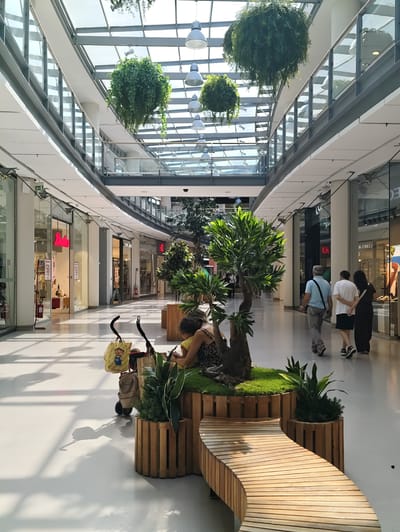 The image shows the interior of a modern shopping mall in Nola, Italy, with a glass ceiling that allows natural light to enter. Hanging plants are suspended from the ceiling, adding a touch of greenery to the environment. In the foreground, a woman with dark hair is sitting on a wooden bench, facing away from the camera, with her back to a stroller. The stroller contains several shopping bags. Further down the mall, a man in a light blue shirt and grey pants, a young person in a white t-shirt and black shorts, and a woman in a dark dress are walking away from the camera. Shops with glass storefronts line both sides of the mall, and display items and signage can be seen. The flooring is a light-colored, smooth surface, reflecting the light. The overall atmosphere is clean, bright, and contemporary.