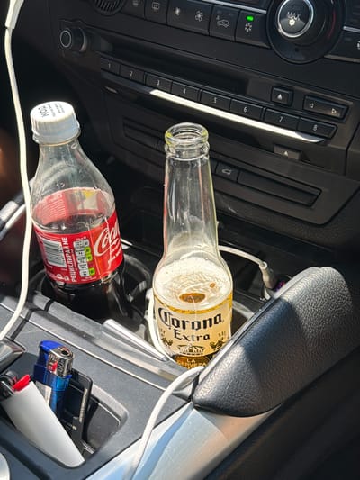 The image depicts the interior of a vehicle's center console area, which appears to be in Peshtera, Bulgaria, based on context not present in the image itself. Two beverages are visible in cup holders: a bottle of Coca-Cola and a bottle of Corona Extra beer, which is partially consumed. A white charging cable is connected to a device and runs towards the back of the center console. Next to the beverages, two lighters are present, one blue and one white. The car's dashboard with climate control buttons and radio controls is visible in the background. The lighting suggests it is daytime, with sunlight creating shadows on the interior surfaces.