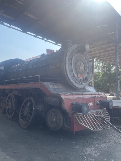 The image shows a vintage steam locomotive, possibly a display piece, situated in a railway structure. The locomotive is painted in maroon and gray, and prominently features the letters "H.E.H" and the number "598." The surrounding environment includes metal framework, suggesting it may be indoors or in a covered area of a railway station. It is difficult to determine the exact location within Anajpur, India without additional context. No people or actions are visible in the photograph. The locomotive sits stationary on a gray surface, possibly concrete or asphalt.