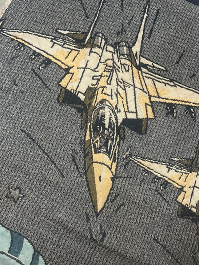 The image shows a close-up view of a woven tapestry or blanket depicting two fighter jets in flight against a dark gray, textured background. The jets are rendered in a stylized, graphic manner with yellow, white, black, and dark green colors. The lead jet is larger and more detailed, showing a cockpit with two figures visible inside. Its nose is pointed and it has prominent wings and tail fins. A second, smaller jet is visible to the right, partially obscured. The background has faint lines that could represent contrails or a sky pattern. There is a small star-like emblem on the left side of the tapestry. The tapestry appears to be made of polyester or a similar synthetic fiber. The image does not contain any elements that would allow for determining its location in Nizhniye Mulli, Russia.