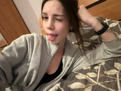 A young woman lying on a patterned bedsheet, wearing a grey zipper hoodie over a black top, playfully sticks out her tongue and winks. Her dark hair falls around her face and onto her arm, which is adorned with a black fitness tracker. The scene is lit by a soft, warm light, casting shadows that subtly highlight the textures of the fabric and her features. A wooden cabinet is visible in the background.