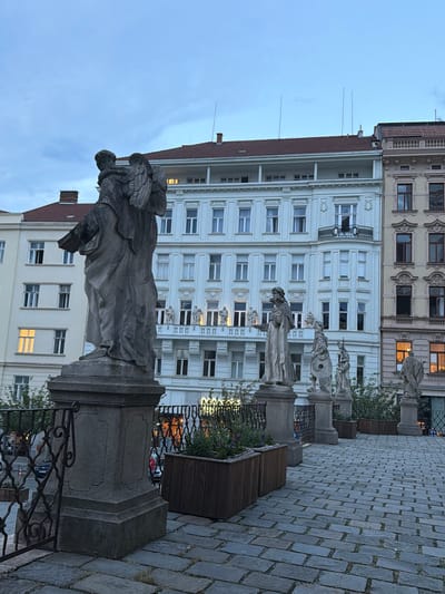 The image depicts an outdoor scene in the city of Brno, Czechia, likely during dusk. In the foreground, a paved walkway made of cobblestones leads towards a row of stone statues situated on pedestals. The statues are positioned along a railing. Behind the statues, several buildings are visible. The central building is a tall, white structure with multiple floors and numerous windows. To its right, another building displays a more ornate facade with yellow and cream-colored bricks and decorative elements around the windows. The sky above is a pale blue, suggesting either early morning or late evening light. Planters with greenery are placed near the statues, and a black metal fence runs along the edge of the walkway. A few cars and people are faintly visible in the background, suggesting an urban environment.