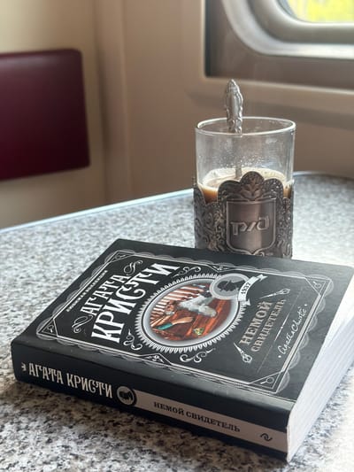 A close-up, high-angle shot captures a book by Agatha Christie and a glass of coffee on a speckled gray surface. The book, titled "Agatha Christie" and "The Silent Witness" in Russian, lies open, displaying an illustration of a person in historical attire. The glass, encased in an ornate metal holder with the Cyrillic letters "РЖД," contains a light brown liquid with a spoon. The background reveals a train compartment with a window and a muted red seat. The scene is devoid of any individuals, suggesting a moment of solitary reading. The overall lighting is natural, coming from the window, casting soft shadows. No specific city or location is identified in the image or its context.