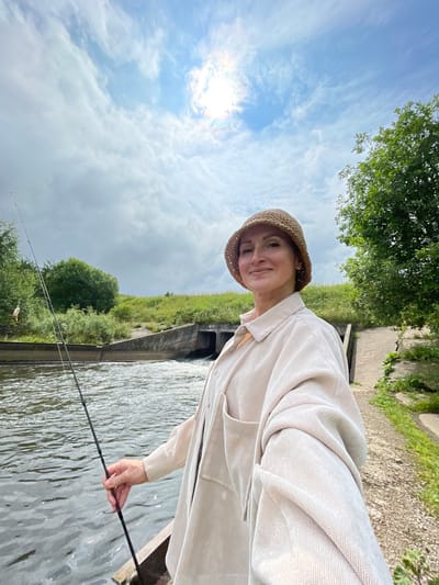 A woman is taking a selfie while fishing on a sunny day. She is wearing a light-colored shirt and a straw hat. She is holding a fishing rod in her right hand. The background shows a river with a small dam or spillway and lush green vegetation. The sky is blue with scattered clouds. The location is the city of Bereznik, Russia.