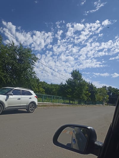 The image shows a view from inside a vehicle, looking out at a street scene. A white SUV is parked on the left side of the road. In the background, there is a line of trees and a green fence. The sky is blue with scattered white clouds. A bird is visible in the sky in the upper right corner of the image. The location is Izberbash, Russia.