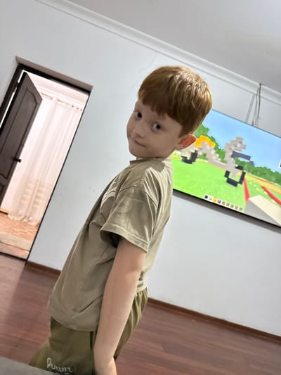 A young boy with light brown hair and blue eyes is standing in a room, looking towards the camera. He is wearing a light brown t-shirt and olive green shorts. The room has white walls and wooden flooring. To his right, a large flat-screen television displays a game that appears to be Minecraft, with pixelated graphics of green terrain and blocky structures. To his left and slightly below him, a doorway is visible, with the door slightly ajar, revealing blinds behind it. The lighting in the room suggests it is daytime, but the specific time and weather conditions are not discernible. The boy's expression is neutral, and he seems to be posing for the photo. The text "FUTURE" is visible on the boy's shorts.