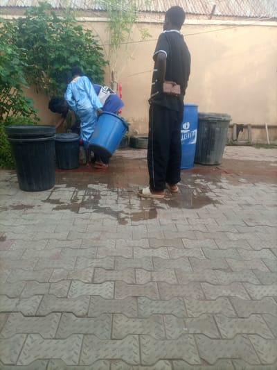 In Wuro Dole, Nigeria, a scene unfolds with several individuals engaged in activity around large containers. A person in a bright blue garment, possibly traditional attire, is bending down and interacting with a dark-colored trash bin. Beside them, another person, partially obscured by the blue-clad individual, is also engaged in the same task. A large blue plastic drum, with the word "NAMA" visible on its side, stands prominently nearby. To the right of the blue drum is a large black trash bin. Further to the right, another tall, dark trash bin is visible. The ground is paved with interlocking bricks, and patches of water are present, suggesting recent rain or cleaning. In the background, a wall with a barbed-wire fence runs along the top, and some greenery is visible, including a leafy bush and a tall, thin plant. The overall impression is one of outdoor domestic or communal work in a semi-urban environment.