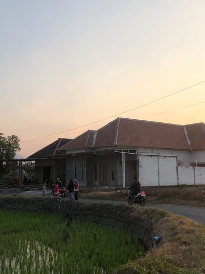 In the city of Sentul, Indonesia, a rural scene unfolds at dusk. The sky transitions from a pale blue to a soft orange, casting a warm glow over the landscape. In the foreground, a lush green rice paddy stretches across the frame, its water reflecting the soft light. A low, stone-lined embankment separates the paddy from a dirt road.
Along the road, a group of people are gathered near a motorcycle. A young child stands to the left, while a group of women, some wearing hijabs, are near the motorcycle. A man rides a motorcycle away from the viewer, his taillight illuminated.
Behind the road, a cluster of houses under construction are visible. They feature red tile roofs and concrete walls, with some sections of white cinder block. Trees and vegetation frame the houses, and in the distance, a hint of mountains can be seen against the horizon. The overall atmosphere is one of peaceful, everyday life in a rural Indonesian setting.