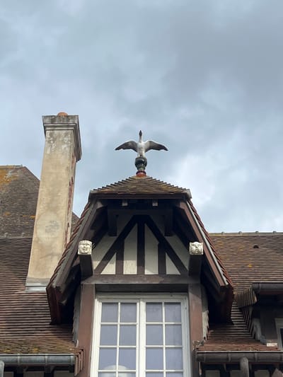 The image captures a portion of a building's rooftop in Deauville, France, under a cloudy sky. Prominently featured is a weather vane in the shape of a swan with outstretched wings, positioned atop a finial on a dormer window. The dormer has a steeply pitched roof with dark wooden beams and white infill, and two grotesque faces carved into corbels protrude from its eaves. To the left of the dormer is a tall, weathered stone chimney with decorative red markings. The surrounding roofs are covered in terracotta tiles.