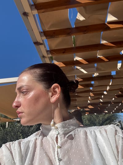 A female individual is depicted in a close-up, profile view, facing left, positioned outdoors under a pergola structure within the Anavissos Municipal Unit, Greece. Her dark hair is pulled back into a bun. She wears a sheer white, high-collared blouse with a textured dotted pattern, fastened with light-colored buttons down the front placket. Her right ear is adorned with a gold-colored ear cuff on the upper helix and a long dangle earring featuring a thin gold-colored bar and a chain descending to a large, irregular, light-colored pearl or stone. The overhead pergola structure comprises white horizontal beams and darker wooden slats partially covered by tan fabric sheets, through which a clear blue sky is visible. Out-of-focus green foliage is present in the lower background. Strong, direct sunlight illuminates the scene.