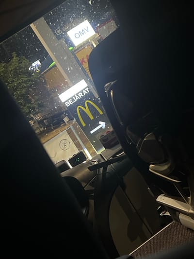 The image captures a view from inside a vehicle at night, likely at a McDonald's drive-thru. The scene is illuminated by artificial lights. A prominent McDonald's sign with the golden arches and the Hungarian word "BEJÁRAT" (entrance) is visible. Above it, an OMV gas station sign is also present. The vehicle's interior, including part of the steering wheel and dashboard, is visible in the foreground, suggesting the photo was taken by a passenger or the driver. The windshield is speckled with what appear to be water droplets or dirt, indicating it might be raining or has recently rained. The environment appears to be an urban setting in Szigetszentmiklós, Hungary, as suggested by the Hungarian text on the McDonald's sign. No individuals are clearly depicted within the scene, but the context implies the presence of at least one person inside the vehicle. The overall atmosphere is dark, with a focus on the illuminated commercial signs.