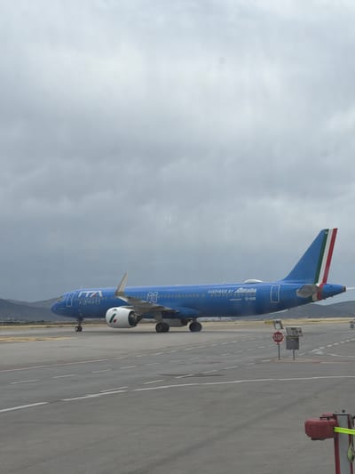 The image captures an ITA Airways Airbus A320 aircraft, registration number EI-HXB, on the tarmac at what appears to be an airport. The plane is predominantly blue with the "ITA Airways" logo prominently displayed. The tail fin features the Italian tricolor. "INSPIRED BY Alitalia" is visible on the fuselage. A "STOP" sign and another indistinct sign are next to the plane on the tarmac. A ground services vehicle is on the right side of the plane, near the rear. The sky is overcast, and a mountain range can be seen in the distance. The airport is within the city of Spata-Loutsa Municipal Unit, Greece.