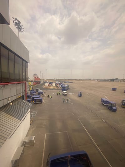The image shows an airport tarmac at Sardar Vallabhbhai Patel International Airport in Ahmedabad, India. The scene is viewed from an elevated perspective, likely from inside the terminal building, looking out through a large window. In the foreground, a portion of the airport building's exterior is visible on the left, with large glass windows and a modern facade. Below the window, a white and grey structure with a corrugated metal roof is also visible.
The tarmac area is busy with various airport operations. Several blue baggage carts, some with the "IndiGo" logo, are present, along with a few white vans and what appear to be small electric tugs used for moving luggage. A few individuals wearing yellow vests and green shirts, likely airport personnel, are seen walking and working on the tarmac. In the middle ground, a red and white control tower stands out, with power lines and other airport infrastructure extending into the distance. The sky is overcast with scattered clouds, and the overall atmosphere suggests a typical day at a large airport. The image captures a snapshot of the operational side of the airport, showcasing the movement of ground support equipment and personnel.
