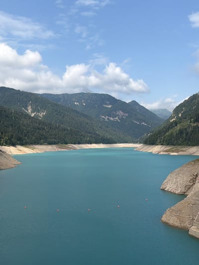 The image depicts a wide, turquoise lake surrounded by lush, tree-covered mountains. The sky above is a bright blue with scattered white clouds. In the foreground, the calm water stretches towards the distant mountains. Several small, orange buoys are visible on the surface of the lake, marking a section of it. The shoreline is characterized by rocky, eroded slopes, with some exposed dry land indicating a lower water level. On the right side of the image, a section of a concrete structure is visible along the bank. The overall scene is a serene natural landscape. The location is the Ampezzo/Dimpeç area in Italy.