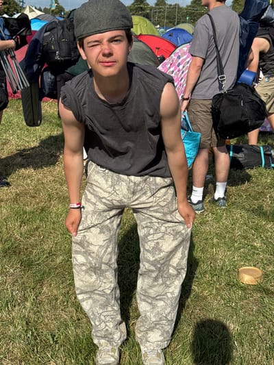 A young man lies on the grass wearing camouflage pants, a gray t-shirt, and a cap. Behind him are tents and other people. Some people are sitting and wearing backpacks. A straw hat sits on the grass in the background. The image is taken from an elevated angle.