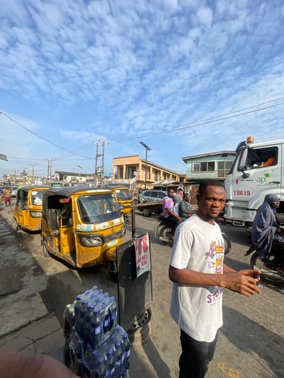 The image depicts a street scene in Akowonjo, Nigeria, under a bright, cloudy sky. In the foreground, a man wearing a white t-shirt with a graphic print and black pants stands facing the camera, holding a dark object in his right hand. To his left, a stack of bottled drinks is visible. Behind the man, several yellow auto-rickshaws are parked along the side of the road. A white truck with "AP" and "ardova plc" branding is also present. Further in the background, various buildings and more vehicles, including a motorcycle with passengers, can be seen. The environment appears to be a busy urban area with overhead electrical wires and streetlights.