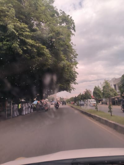 The image captures a street scene in Meunasahtimu, Indonesia, viewed from inside a moving vehicle, with part of the dashboard visible at the bottom. The sky is overcast with light grey clouds, suggesting either an early morning, late afternoon, or simply a cloudy day.
On the left side of the road, a dense canopy of large green trees provides significant shade. Beneath these trees, a gathering of people is visible. Many individuals, predominantly women, are dressed in long white robes and white head coverings, resembling uniforms or religious attire. Several motorbikes are parked or slowly moving among them, and a few men are also present. They appear to be congregating or waiting by the roadside.
The road itself is asphalt, featuring a median strip with a low curb and sparse vegetation. On the right side of the road, a row of smaller, neatly trimmed trees lines the thoroughfare. Beyond these trees, various buildings, characteristic of urban or semi-urban architecture, can be seen in the background. Traffic, including cars and motorbikes, is observed moving in both directions on the road. No specific text is clearly legible within the image. The overall scene depicts a busy local street environment with a distinct cultural element due to the attire of the gathered people.