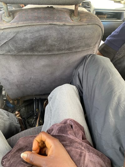 The image depicts the interior of a vehicle, viewed from a passenger's perspective in Unguwar Gaya, Nigeria. The upper half of the frame is dominated by the worn, grey fabric back of a car seat, including its metal headrest support rods. Below the seat, in the footwell, a black bag with a visible gold zipper and other indistinct objects are present. In the lower foreground, the left leg of the individual holding the camera, clad in light grey denim, is visible. Their right hand, with glitter nail polish, rests on a folded brown corduroy garment. Adjacent to the photographer's leg, to the right, is the right leg of another passenger, covered in dark grey, possibly nylon, material. A partial view of a third individual's navy blue long-sleeved arm is visible to the right of the front seatback.