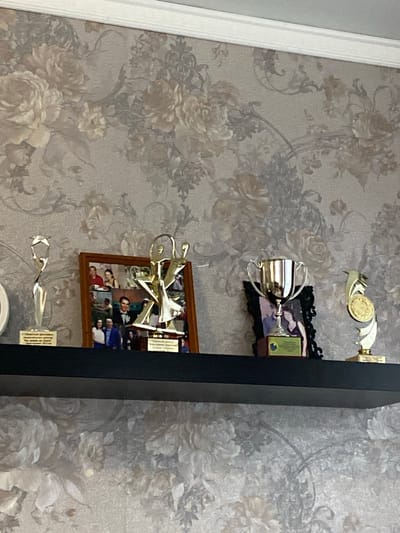 A shelf with awards and framed photographs is mounted on a wall with floral wallpaper. From left to right, the shelf holds a silver abstract trophy with a star on top, a framed collage of photographs, a gold abstract trophy, a silver trophy cup with handles, and a gold trophy with a circular medallion. The photograph collage features several pictures of people in various settings, including formal portraits and outdoor scenes. The trophies appear to be awards for achievements or competitions. The overall setting suggests a display of personal accomplishments or memories.