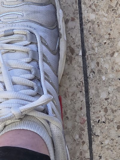 A close-up, high-angle view depicts an individual's left foot, stationary on a light-colored, speckled terrazzo or aggregate concrete surface in Eeklo, Belgium. The foot is clad in a worn, light gray and white sneaker constructed with mesh and synthetic overlays. Light gray laces are tied at the shoe's vamp. A small red detail is visible on the inner side of the shoe, near the sole. Above the shoe's collar, a segment of bare skin and a black garment, consistent with a sock or trouser cuff, are visible. To the immediate right of the shoe, a distinct dark linear seam or grout line bisects the ground surface vertically.