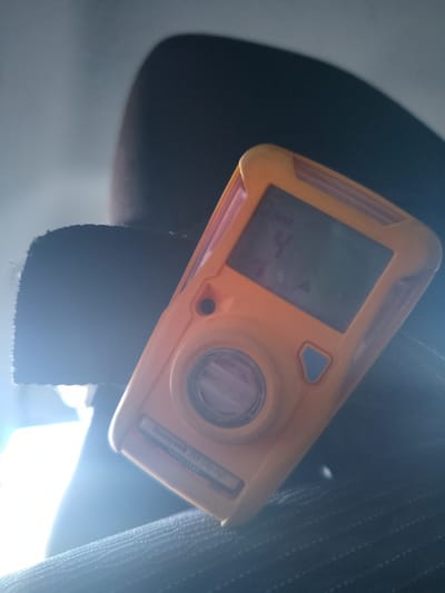 An orange Honeywell BW Clip H2S gas detector is positioned on a dark, textured fabric surface, possibly a piece of clothing or upholstery. The device's screen displays "4" with a triangle and "H2S" below it, indicating a reading of 4 parts per million of hydrogen sulfide. The background is out of focus and appears to be a light grey surface. A bright light source is visible from the left, casting a lens flare and highlighting the fabric and the gas detector. The location is specified as Жарсуат ауылдық округі, Kazakhstan.