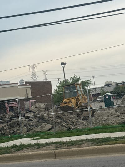 The image depicts a construction site in Niles, Illinois, USA. Two heavy machinery vehicles, a yellow bulldozer and a pink excavator, are present. The bulldozer is positioned in the center right of the frame, partially obscured by debris. The pink excavator is located to the left, also amidst piles of dirt and rubble. A green portable toilet is situated to the right of the bulldozer. In the background, several large electrical towers are visible against an overcast sky, and buildings suggest an urban or suburban setting. A chain-link fence runs across the foreground, separating the construction area from the street. The ground in front of the fence is covered with grass and a concrete path.