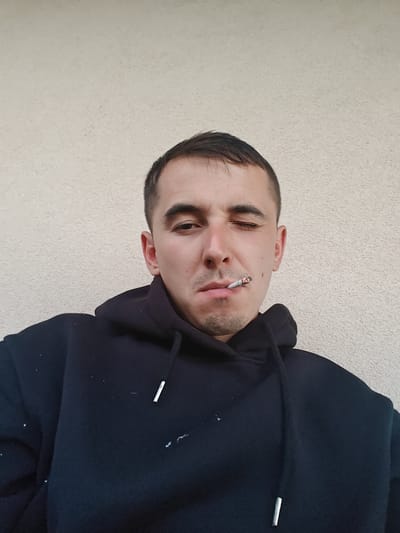 A close-up, medium shot shows a man with short, dark hair and a light complexion. He is wearing a black hooded sweatshirt. The man is looking directly at the camera, with his left eye partially closed. A lit cigarette is held in his mouth, between his lips. He has stubble on his chin. The background is a plain, textured wall. The image appears to be taken outdoors, with no other people or objects visible in the frame. The scene is located in the city of Shkurupiivka, Ukraine.