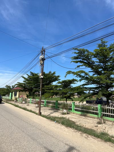 The image depicts a street scene in Dalam, Indonesia, under a bright, clear blue sky, suggesting a sunny day. The perspective is from the side of a road, looking upwards towards a tangle of overhead power lines crisscrossing the frame. A wooden utility pole, laden with these wires and various electrical components, stands prominently on the left side of the image.

Flanking the road is a dirt path, bordered by a white metal fence with green accents. Beyond the fence, lush green trees with dense foliage are visible, casting shadows on the ground. In the background, a building with a yellow facade and a red roof can be seen, along with more sections of the distinctive green and white fence. The presence of the fence and the building suggests a residential or possibly a public area.

The road surface itself appears to be asphalt, textured with small stones. The overall impression is one of a typical day in a moderately developed area, with infrastructure like power lines coexisting with natural elements like trees and clear skies. No people are visible in the image, and the scene appears to be static.