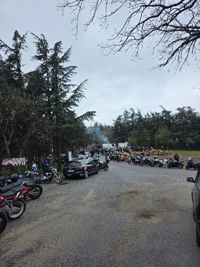 This image captures an outdoor gathering of individuals and vehicles in Chargan, Bulgaria, under an overcast, grey sky. The scene is dominated by a grey, paved surface in the foreground and center, showing signs of wear and minor staining.

Numerous motorcycles are present, parked in various configurations. Several are positioned diagonally in the left foreground, some with helmets resting on their seats. Further into the mid-ground, a dark-colored SUV is parked on the paved surface, facing away from the viewer, with a Bulgarian license plate visible. Many more motorcycles are clustered on both the paved area and extending into a grassy or dirt field on the right.

A large number of people are scattered throughout the mid-ground and background, many appearing to be wearing motorcycle apparel. They are standing and conversing in groups. In the far background, several white, temporary structures are visible, with a plume of white smoke rising from their vicinity. To the right of the main paved area, multiple wooden picnic tables with attached benches are arranged on the grassy terrain, some with individuals seated or standing nearby.

The environment features dense, dark green coniferous trees lining the left side and extending into the background. Bare deciduous tree branches frame the top portion of the sky. On the far left, a banner or sign with graffiti-style lettering, including the word "SOFIA," is attached to a tree. A partial view of a dark vehicle is visible on the extreme right edge of the frame.