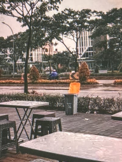 The image depicts an outdoor scene with a clear focus on a small seating area, suggesting a commercial establishment. In the foreground, a square, light-colored table is visible, positioned on a brick-patterned surface. To the left of this table are two dark, woven-style stools. To the right, another similar stool is partially visible. In the mid-ground, a larger, dark trash bin with an orange lid stands prominently.

Behind the trash bin, a paved walkway or street is observed, with a person in a blue garment seated on a scooter or motorcycle. The background reveals a landscaped area with various trees and shrubs, interspersed with buildings that appear to be modern, multi-story structures with a light-colored facade and dark window frames. A street light with a curved pole is also noticeable in the left background. The overall impression is of a contemporary urban environment, possibly a cafe or restaurant patio in a city like Babahjurong, Indonesia, given the context provided. The image quality is somewhat grainy, suggesting it might be an older photograph or taken with a lower-resolution device.