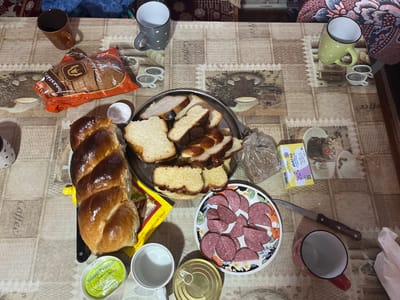 This is a top-down, medium shot of a table set for a meal in Pivniceni, Moldova. The table is covered with a beige tablecloth featuring a repeating pattern of coffee cups and saucers, and lace trim. On the table, there is a braided loaf of sweet bread, a package of sliced bread, a plate of sliced sausage, a small dish of what appears to be butter or spread, and a tin can. Several mugs and cups are also visible, including a brown mug, a gray polka-dot mug, a green polka-dot mug, a white mug, and a red mug. A knife is placed next to the plate of sausage. The scene appears to be indoors, likely in a home, given the casual setting and the presence of personal items like mugs. There are no people visible in the image, and the focus is on the food and table setting.