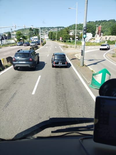 The image depicts a sunny day on a multi-lane road in Sotteville-lès-Rouen, France. The scene is dominated by vehicles and road infrastructure. In the foreground, the dashboard and windshield of a vehicle are visible, with a GPS device mounted on the right side.

Two cars are prominently featured on the road ahead. The car on the left is a dark grey Dacia SUV, with the license plate "GH 022 LO" clearly visible. To its right, a black classic Mini Cooper is driving in the same direction, displaying the license plate "BP 320 NZ". Further ahead, a white SUV is turning right on a separate lane. The road appears to be an approach to an intersection or a complex interchange, indicated by multiple lanes, traffic lights, and concrete barriers.

In the background, lush green hills rise, dotted with buildings, suggesting a semi-urban or suburban environment. A cement factory or construction site with industrial machinery is visible on the right side of the road. The sky is clear and blue, characteristic of a bright, fair-weather day, likely mid-morning or afternoon given the shadows. The overall impression is one of typical daily traffic flow in a French town. No people are clearly discernible within the vehicles. The visible text includes license plates and signage on the industrial buildings.