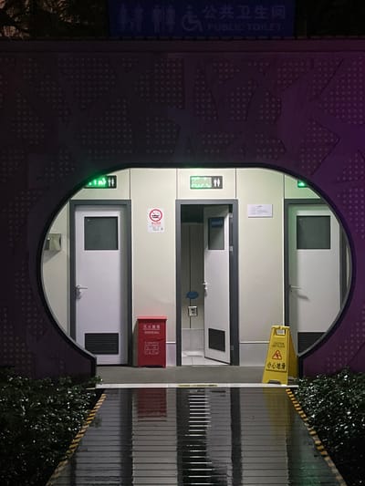 An exterior nighttime view in Yanqiao, China, depicts a public toilet facility enclosed within a dark, decorative archway featuring a circular opening. Above the arch, a blue sign with white pictograms (male, female, disabled) and text "公共卫生间 PUBLIC TOILET" is visible.
Inside the circular opening, three individual toilet doors are arranged. All doors are white with dark rectangular windows at the top and louvered vents at the bottom.
* The leftmost door is closed, with a green illuminated "occupied" indicator sign above it.
* The central door is slightly ajar, revealing a tiled interior, also with a green illuminated "occupied" indicator sign above it.
* The rightmost door is closed, with a green illuminated indicator sign partially visible above it.
Between the leftmost and central doors, a white "No Smoking" sign is mounted on the wall above a red fire extinguisher cabinet. Between the central and rightmost doors, a small white sign with unreadable text is affixed to the wall. A yellow A-frame "CAUTION WET FLOOR 小心地滑" sign stands near the rightmost door.
The ground directly in front of the doors is a dark, smooth surface. A reflective, wet pathway with horizontal grooves extends from the facility towards the viewer, showing reflections of the structure and internal lights. Dark green shrubbery borders the pathway on both sides. The scene is illuminated by artificial lighting, indicating nighttime conditions. No individuals are present.