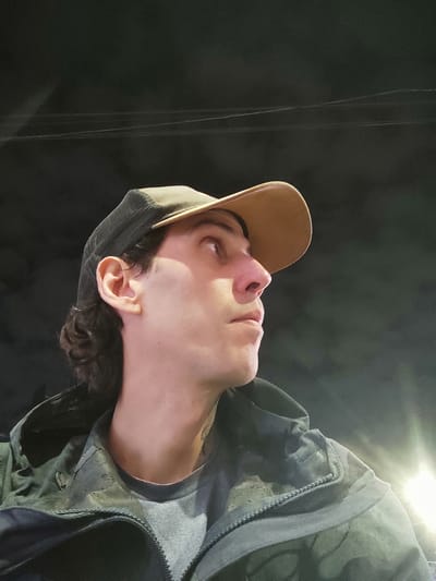A close-up, angled shot captures a person, likely male, in three-quarter profile looking downwards and to their right. The person has dark, curly hair visible under a dark, mesh-back cap with a contrasting light brown brim. They are wearing a dark green or grey hooded jacket over a lighter grey t-shirt. A small hoop earring is visible in their right ear.
The background is predominantly dark and out of focus, suggesting a nighttime or low-light indoor setting. A bright, artificial light source is reflecting strongly in the lower left portion of the image. Faint, thin, parallel vertical lines are discernible in the dark background, possibly indicating wires, strings, or light streaks.
The person's expression appears focused or contemplative as they gaze downwards. Details of their clothing include a visible zipper on the jacket. No text is visible in the image. The location context is Lanús Este, Argentina.