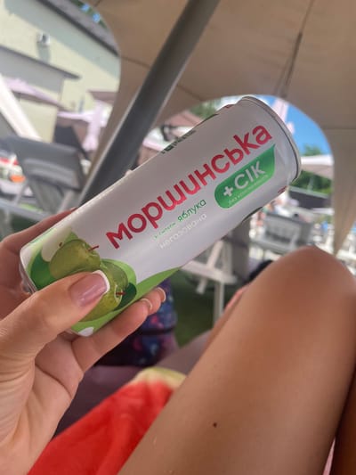 A person's hand with light skin and a French manicure is holding a white and green aluminum can of "МОРШИНСЬКА + Сіk" (Morshynska + Juice) with images of green apples. The text on the can indicates it is apple-flavored and "без консервантів" (without preservatives). The can is angled towards the viewer and is partially obscuring a person's bare leg. In the background, there are blurry elements suggesting an outdoor setting, possibly a patio or poolside area, with furniture and some vegetation. The environment appears to be sunny. The context provided indicates this scene is in Zazymia, Ukraine.