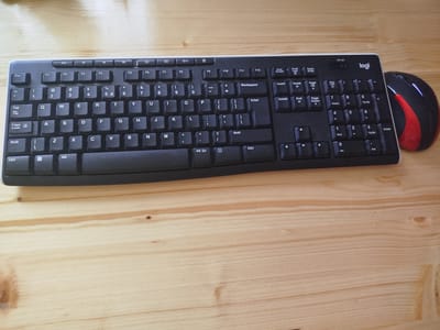 The image shows a black Logitech keyboard and a red and black computer mouse placed on a light-colored wooden desk. The keyboard is a full-sized QWERTY keyboard with a numeric keypad. The mouse is positioned to the right of the keyboard. The wooden desk has a natural wood grain pattern. There are no people or other objects visible in the scene. The image is a top-down view of the desk. The location of Cēsis, Latvia is not ascertainable from the provided image.