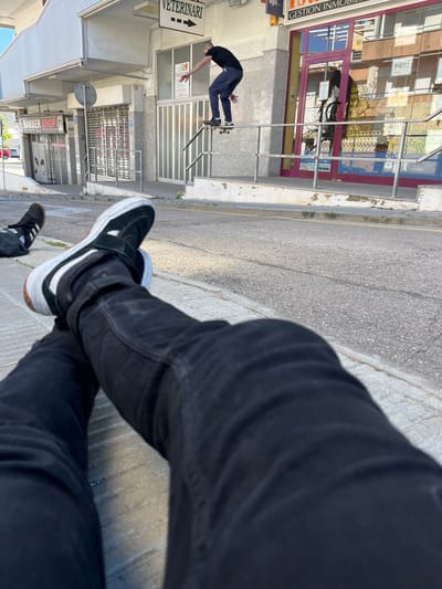 A street scene in Calafell, Spain, is captured from a low vantage point. The foreground features the crossed legs of an individual wearing black jeans and black-and-white skateboarding shoes, occupying the right side of the frame. In the background, a man is visible on a skateboard, positioned in front of a building that houses businesses including a veterinary clinic and a barber shop; signs for each are present. Further down the street, another building, a real estate office ("Gestion Inmobiliaria"), can be seen. The street itself is paved with asphalt, and the overall setting is an urban environment.