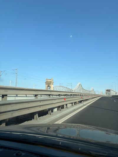 The image depicts a forward-facing perspective from inside a moving vehicle on a multi-lane highway bridge in the Cernavodă/Borcea area, Romania. A clear, bright blue sky with no clouds dominates the upper portion, featuring two faint circular artifacts in the upper right. The lower foreground shows the dark, reflective surface of the vehicle's dashboard. A multi-tiered metal guardrail system, equipped with red triangular reflectors, runs along the left side, separating the highway from an adjacent railway line, indicated by overhead catenary wires and support poles. The asphalt highway surface, marked with white lines, extends into the distance, curving gently to the right. In the background, the ornate stone tower and extensive steel trusswork of the historic Anghel Saligny railway bridge are prominent, running parallel to the highway bridge structures. Further to the right, additional modern bridge sections are visible. No human figures or other vehicles are present on the depicted highway segment.