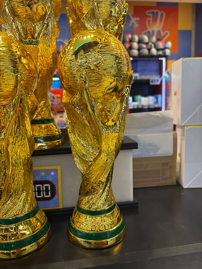 The image shows a close-up of several golden trophies, resembling the FIFA World Cup trophy, displayed on a black shelf. The trophy in the foreground is the most detailed, featuring a globe at the top and intricately sculpted figures holding it. The base of this trophy has green bands and appears to have text inscribed, though it is partially obscured. To the left of this trophy, another similar trophy is visible, also with green bands. In the background, out of focus, there are shelves stocked with various items, possibly in a retail setting. On the far right, stacks of white boxes are present. A digital display with the number "100" is visible on a colorful sign in the lower left portion of the image, with stars above it. The overall lighting suggests an indoor environment. The location is identified as Aeropuerto Internacional Ezeiza, Argentina, indicating this is likely a souvenir shop or display within the airport. There are no people visible in the image.