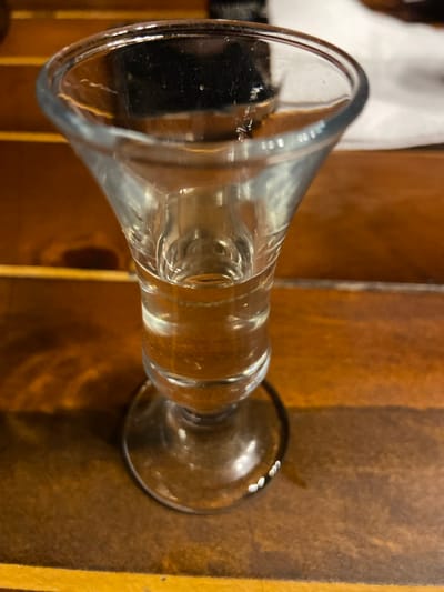 A clear glass shot glass, containing a transparent, colorless liquid filling approximately one-third of its volume, rests on a polished, warm-brown wooden surface in Dulovine, Montenegro. The shot glass features a flared rim, a tapered body, and a wide circular base. Visible reflections on the glass and liquid indicate their transparent properties. Minor smudges and dust are present on the glass surface, and small white points of light are visible along the edge of the glass's base. The wooden surface, composed of multiple planks, exhibits distinct grain patterns. The background is out of focus, showing additional portions of the wooden surface and indistinct elements. No individuals or specific actions are discernible within the frame.