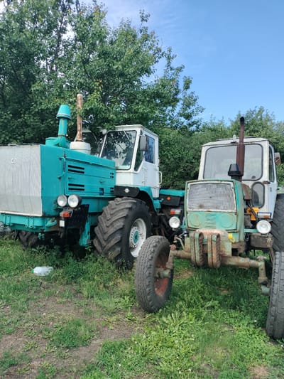 This image shows two old, weathered tractors parked in a grassy field in Stanychne, Ukraine. The tractor in the foreground is a light green color, showing significant signs of rust and wear. It has a prominent, mesh-like grille and large, thick tires. The second tractor, to the left and slightly behind, is primarily turquoise and white. It appears larger and somewhat better maintained, though still aged. Both tractors have tall exhaust pipes.
The scene is set outdoors, with lush green trees and foliage forming a dense backdrop behind the tractors. The ground is covered in grass and some patches of bare earth, with weeds and wildflowers visible. In the lower left quadrant of the image, a white plastic bottle with a green cap lies on the grass.
The sky above is a clear, bright blue, suggesting it is daytime, likely late morning or early afternoon given the lighting. There are no people visible in the image. The overall impression is one of abandonment or disuse, with the tractors appearing to be stationary, perhaps no longer in active service. The rust on the green tractor and the slightly faded paint on the turquoise one evoke a sense of nostalgia and the passage of time. The image is an outdoor photograph, taken from a medium distance, capturing the entirety of both machines and their surroundings. The word "XT3" is visible in white lettering on a metallic rectangular panel on the front of the turquoise tractor.